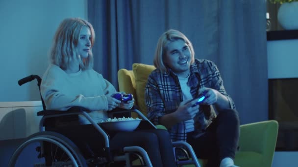 Pareja rubia de hombre y mujer jóvenes en una silla de ruedas sentados en la sala de estar y jugando con joysticks - Imágenes, Vídeo