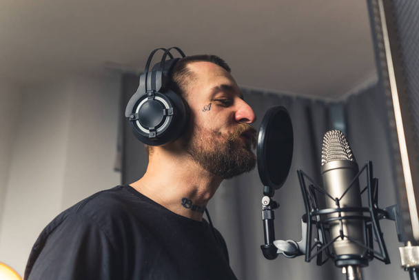 a tattooed good-looking bearded young man with headphones singing in the microphone studio shot medium closeup musical concept profile shot - Photo, Image