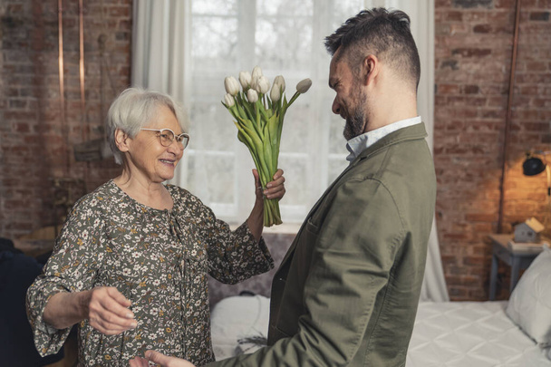 két kaukázusi ember egy modern lakásban ünnepli az Anyák napját, idős nő fehér virágokkal a kezében és középkorú fia - Fotó, kép