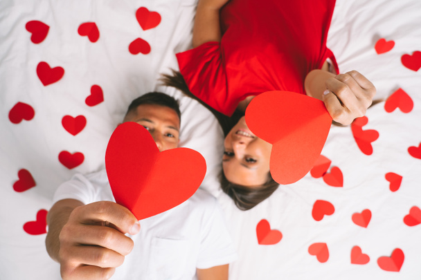 Pohled shora na mladý atraktivní zamilovaný pár ležící na posteli a ukazující srdce z červeného papíru. Šťastný Valentýn koncept - Fotografie, Obrázek