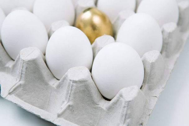 Белые сырые куриные яйца и одинокое золотое яйцо в лотке вид сбоку - Фото, изображение