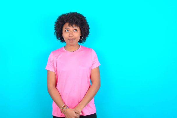 Jeune femme afro-américaine portant un T-shirt rose sur fond bleu regardant de côté dans l'espace vide réfléchi - Photo, image