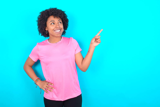 Smiling Young African American woman wearing pink T-shirt over blue background indicating finger empty space showing best low prices - Fotografie, Obrázek