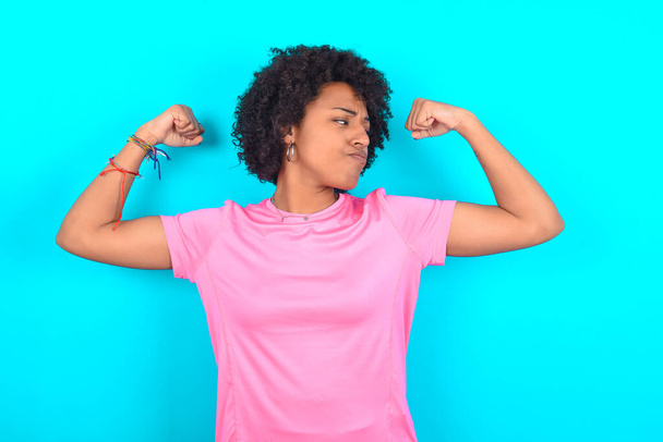 Young African American woman wearing pink T-shirt over blue background showing arms muscles smiling proud. Fitness concept. - Fotoğraf, Görsel