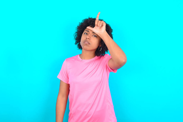 Young African American woman wearing pink T-shirt over blue background making fun of people with fingers on forehead doing loser gesture mocking and insulting. - 写真・画像