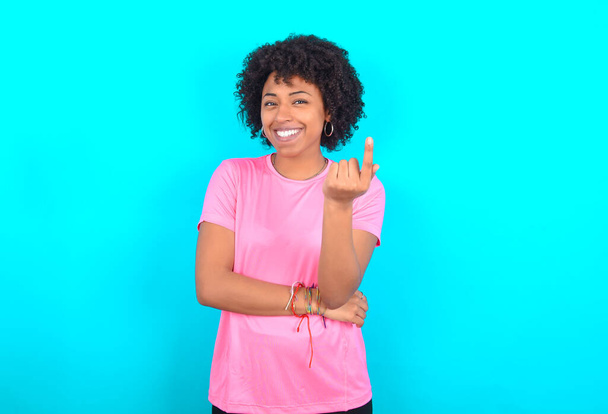 Молодая афроамериканка в розовой футболке на синем фоне, манящая рукой, приходит сюда приветствуя счастливую и улыбающуюся - Фото, изображение