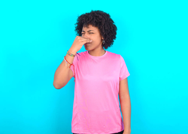 Jonge Afro-Amerikaanse vrouw draagt roze T-shirt over blauwe achtergrond ruikt iets stinkend en walgelijk, ondraaglijk geur, houdt adem met vingers op de neus. Slechte geur - Foto, afbeelding