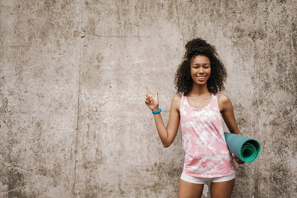 Young black sportswoman smiling and pointing finger upward while standing by concrete wall - Foto, Imagen