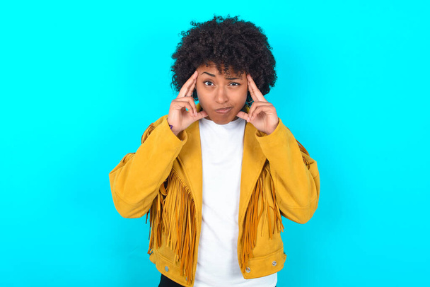 Serious concentrated Young African American woman wearing yellow fringe jacket over blue background keeps fingers on temples, tries to ease tension, gather with thoughts and remember important information for exam - Foto, Imagem