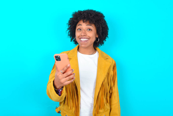 Young African American woman wearing yellow fringe jacket over blue background holds mobile phone in hands and rejoices positive news, uses modern cellular - 写真・画像