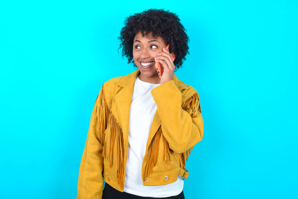Pleasant looking happy Young African American woman wearing yellow fringe jacket over blue background has nice telephone conversation and looks aside, has nice mood and smiles positively while talks via cell phone - Foto, Imagem