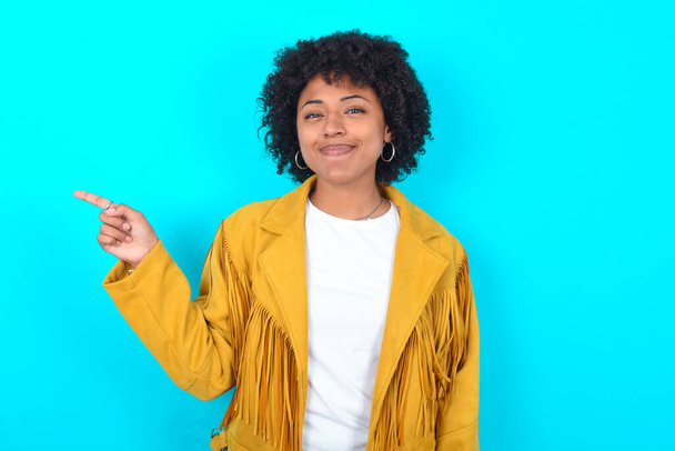 A fiatal afro-amerikai nő sárga rojtos kabátot visel a kék háttér felett, az üres tér egyik oldaláról a másikra mutatja a hirdetést. Emberek és promóciós koncepció - Fotó, kép