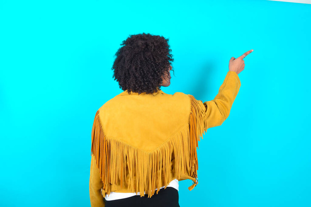 Giovane donna afroamericana indossa giacca frangia gialla su sfondo blu che punta all'oggetto su spazio copia, vista posteriore. Gira le spalle. - Foto, immagini