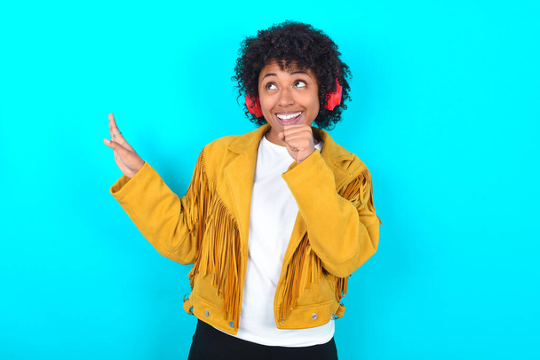 Happy Young African American woman wearing yellow fringe jacket over blue background sings favourite song keeps hand near mouth as if microphone wears wireless headphones, listens music - Fotografie, Obrázek