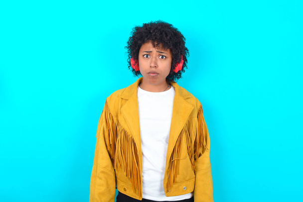 Дуже невдоволена молода афроамериканська жінка, одягнена в жовтий піджак на синьому фоні, спантеличена тим, що вона носить стерео навушники, слухаючи музику під час ходьби на вулиці. - Фото, зображення
