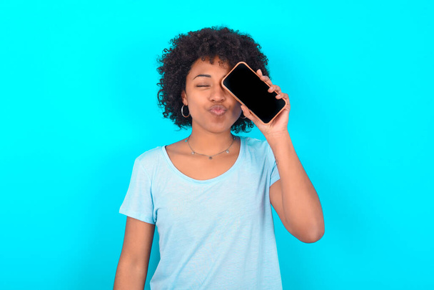 Adorable femme afro-américaine portant un t-shirt bleu sur fond bleu tenant un dispositif moderne couvrant l'œil avec des lèvres boudées - Photo, image