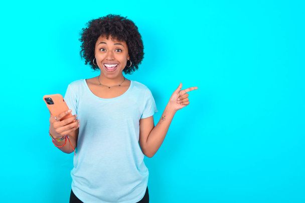 Verbaasde Afro-Amerikaanse vrouw draagt blauw T-shirt over blauwe achtergrond met haar telefoon en wijzend met de vinger opzij naar lege kopieerruimte - Foto, afbeelding