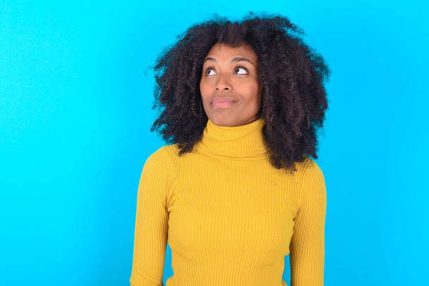 Афроамериканка, одягнена в жовту білизну на синьому фоні, дивиться в порожній простір замислено. - Фото, зображення