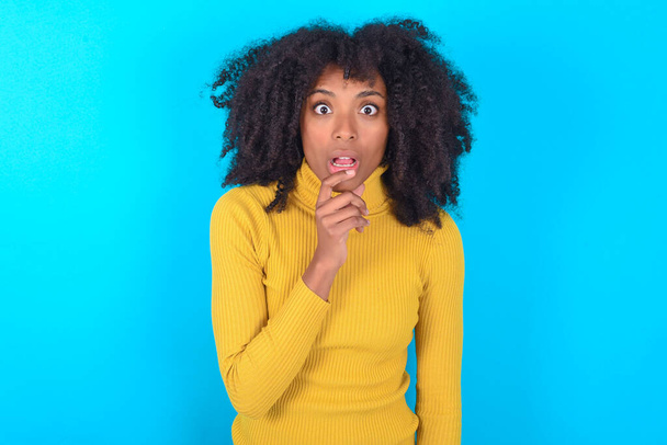 Nervózní zmatený Afroameričanka na sobě žlutý rolák přes modré pozadí otevře ústa od překvapení, reaguje na náhlé zprávy. - Fotografie, Obrázek