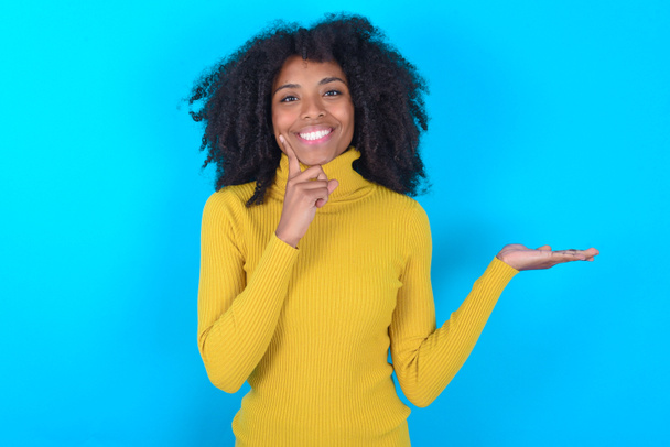 Смешная афроамериканка в желтой водолазке на голубом фоне держит открытую ладонь новый продукт большое предложение - Фото, изображение