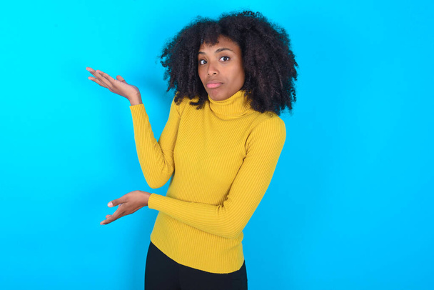アフリカ系アメリカ人の女性は、両手で何か奇妙なことを示していると言って、青い背景の上に黄色のボトルネックを身に着けています:私はこれが何であるかわからない。広告コンセプト. - 写真・画像