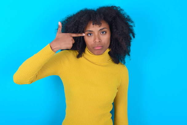 Mujer afroamericana infeliz con cuello alto amarillo sobre fondo azul hace gesto suicida e imita arma con mano, curva labios mantiene dos dedos en la sien, dispara, estar cansado de todo, - Foto, Imagen