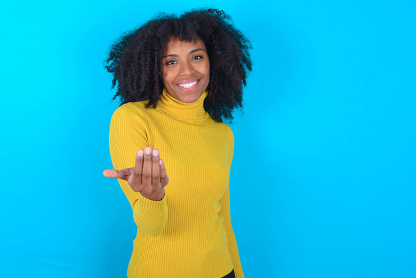Αφρο-Αμερικανίδα που φοράει κίτρινο ζιβάγκο πάνω από μπλε φόντο προσκαλώντας την να έρθει με το χέρι. Χαίρομαι που ήρθες. - Φωτογραφία, εικόνα