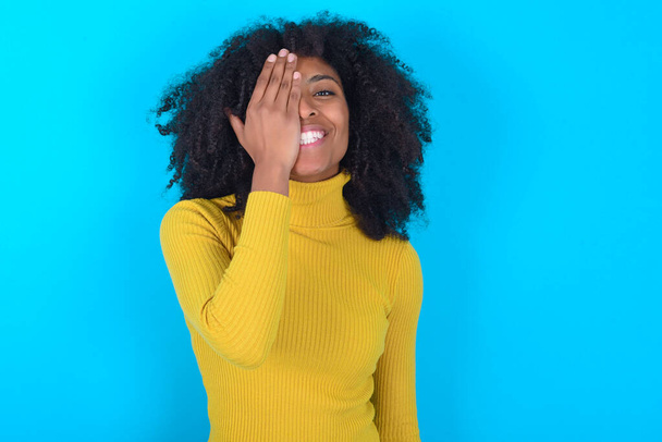 Africano americano mulher vestindo gola alta amarela sobre fundo azul cobrindo um olho com a mão, sorriso confiante no rosto e emoção surpresa. - Foto, Imagem
