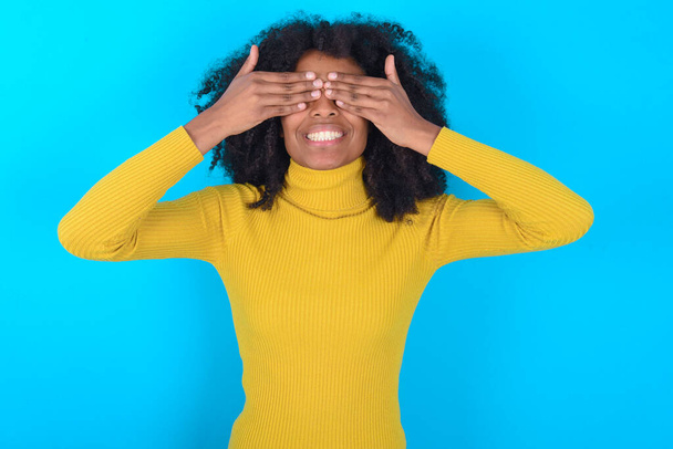 African American woman wearing yellow turtleneck over blue background covering eyes with hands smiling cheerful and funny. Blind concept. - Photo, Image