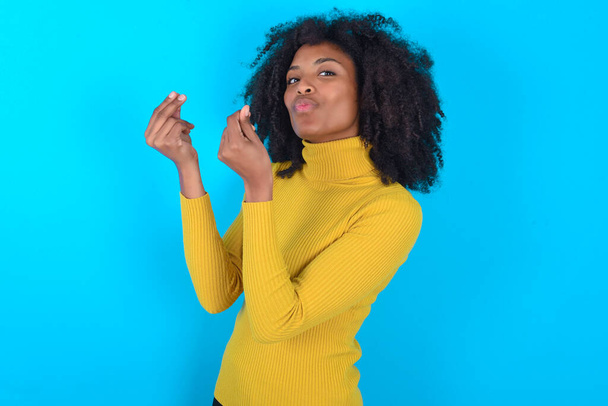 Mujer afroamericana con cuello alto amarillo sobre fondo azul haciendo gesto de dinero con las manos, pidiendo pago de sueldo, negocio millonario - Foto, imagen