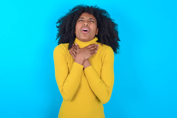 Donna afroamericana con il dolcevita giallo su sfondo blu urlando soffocare perché strangolamento doloroso. Problemi di salute. Asphyxiate e concetto di suicidio. - Foto, immagini