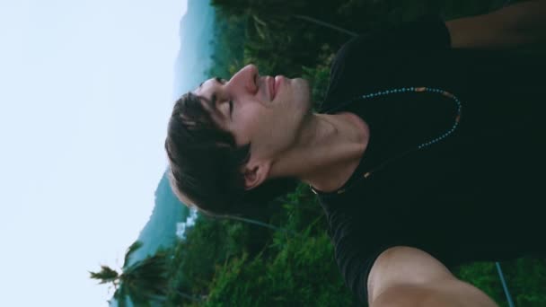 Vertikales Videoporträt eines lächelnden jungen Mannes, der ein Selfie-Video auf grünem Dschungel-Wald und Palmen-Hintergrund macht. Junge männliche Träumer genießen Reisen in tropischem Land - Filmmaterial, Video