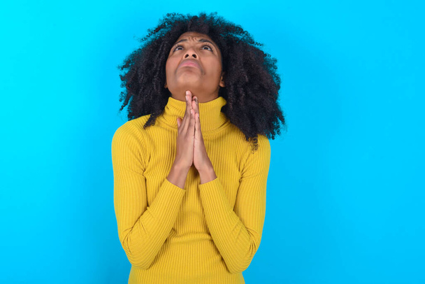 アフリカ系アメリカ人の女性は、青い背景の物乞いと祈りの上に黄色のタートルネックを身に着けていると一緒に顔に希望の表現は非常に感情的かつ心配。神よ - 写真・画像