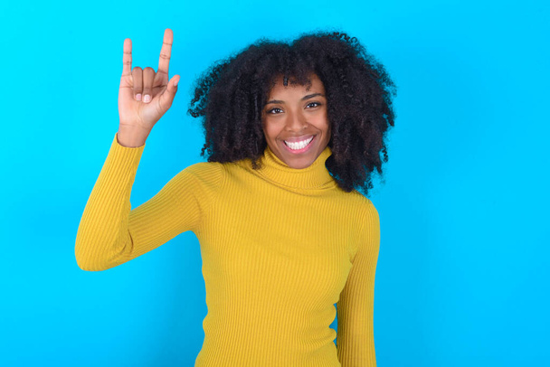Mujer afroamericana con cuello alto amarillo sobre fondo azul haciendo un gesto de roca y sonriendo a la cámara. Listo para ir a su concierto favorito de la banda. - Foto, Imagen
