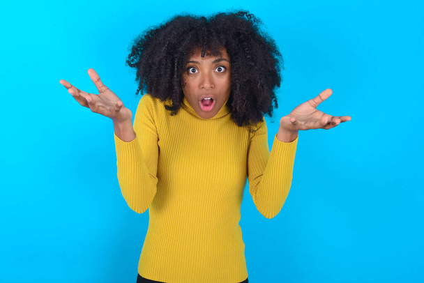 Frusztrált afro-amerikai nő sárga garbót visel a kék háttér úgy érzi, zavaros és tétova, visel vállát zavarodottság, tartja a száját széles körben nyitva, nem tudja, mit tegyen. - Fotó, kép