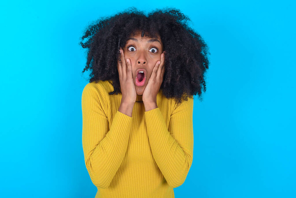 Verängstigte Afroamerikanerin mit gelbem Rollkragen vor blauem Hintergrund schockiert über Preise im Geschäft, Menschen und menschliche Emotionen - Foto, Bild