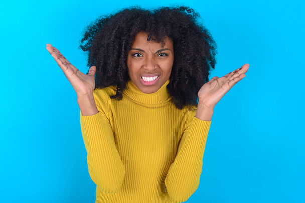 Indignée femme afro-américaine portant col roulé jaune sur fond bleu gestes de perplexité, fronce les sourcils face à l'insatisfaction. - Photo, image