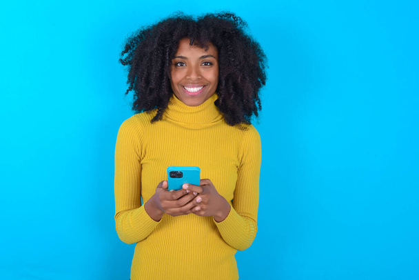 青い背景の上に黄色のボトルネックを身に着けているアフリカ系アメリカ人の女性は、遠くの通信を楽しんで、携帯電話を使用して、サーフィン高速無制限のインターネット、快適な笑顔を持っている、オンラインショッピングになります, - 写真・画像