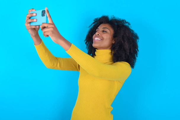 Afroamerykanka ubrana w żółty golf na niebieskim tle robi sobie selfie, aby zamieścić go w mediach społecznościowych lub mieć rozmowę wideo z przyjaciółmi. - Zdjęcie, obraz