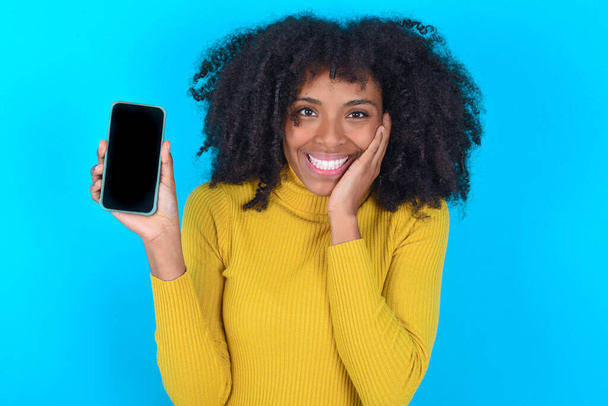Mujer afroamericana con cuello alto amarillo sobre fondo azul de la mano el uso de la tecnología moderna toque la palma de la cara asombrado gritar impresionado wow omg increíble inesperada - Foto, Imagen