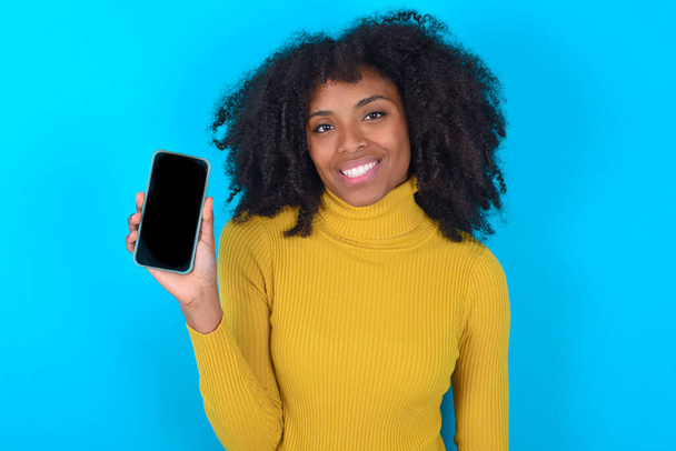Smiling African American woman wearing yellow turtleneck over blue background Mock up copy space. Hold mobile phone with blank empty screen - Fotoğraf, Görsel