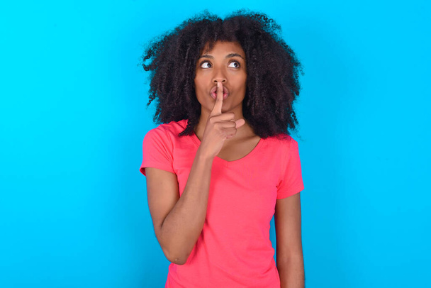 Mavi arka plan sessizlik jesti üzerine pembe tişört giyen Afro-Amerikalı bir kadın işaret parmağını dudaklara doğru tutuyor. Sırrımızı paylaşmamamızı istiyor. - Fotoğraf, Görsel
