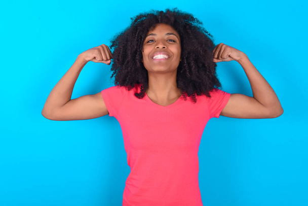 Sterke krachtige Afro-Amerikaanse vrouw draagt roze T-shirt over blauwe achtergrond tandenglimlach, heft armen en toont biceps. Kijk naar mijn spieren.! - Foto, afbeelding