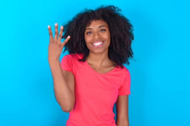 Afrikkalainen amerikkalainen nainen yllään vaaleanpunainen T-paita sininen tausta hymyilevä ja etsivät ystävällinen, osoittaa numero neljä tai neljäs käsi eteenpäin, laskenta alas - Valokuva, kuva