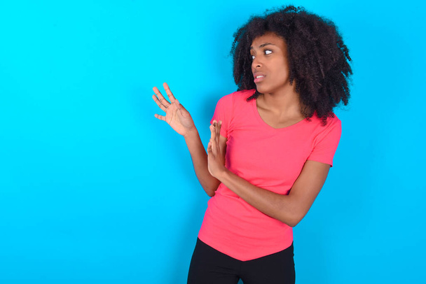 Mavi arka planda pembe tişört giyen hoşnutsuz Afro-Amerikalı kadın ellerini boş alana uzatıyor ve yakına gelmemesini istiyor. - Fotoğraf, Görsel