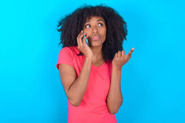 Femme afro-américaine portant un t-shirt rose sur fond bleu parle sur téléphone mobile passe du temps libre à l'intérieur appels à un ami. - Photo, image