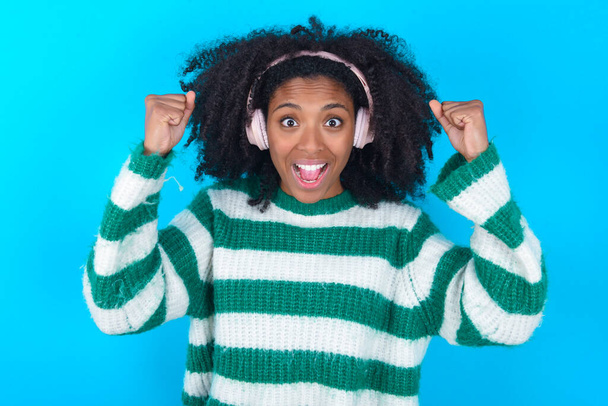 Емоційна афро-американська жінка, одягнена в смугастий светр над синім фоном, голосно вигукує, ніби переможець піднімає стиснуті кулаки, щоб рот був відкритий, носіння стерео навушників на вухах робить жест, слухає улюблену музику - Фото, зображення