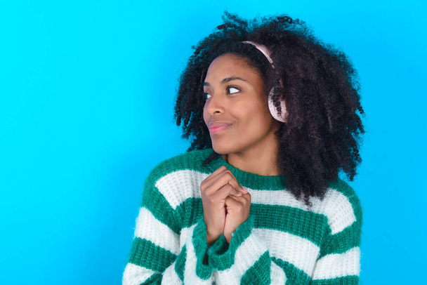 Afroamerikanerin in gestreiftem Pullover vor blauem Hintergrund trägt Stereo-Kopfhörer, hört konzentriert Musik und blickt interessiert zur Seite. - Foto, Bild