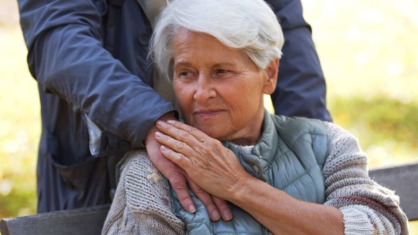 Porträt einer älteren kaukasischen Dame berührt die Hand ihres Mannes, der wegschaut, Mann steht hinter seiner Frau  - Foto, Bild