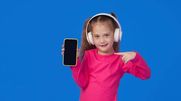 明るいピンクのセーターのかわいい女の子は、彼女の手の中に携帯電話を保持し、無線ヘッドフォンで音楽を聴く。ガジェットへの子供のアプリケーションの概念 - 写真・画像